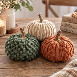 May include: Three decorative pumpkins crafted from knit fabric. One is sage green with a bobble texture, one is cream with a ribbed pattern, and one is burnt orange with a cable knit design. Each pumpkin features a brown, textured stem on top.