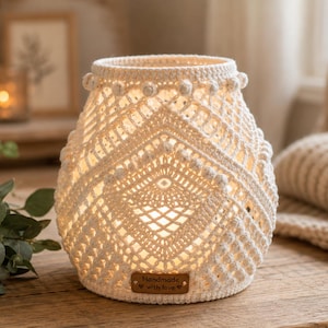 May include: A cream-colored, crocheted lantern cover featuring an intricate diamond pattern and small bobble accents. A rectangular brown leather tag is attached to the base with the text "Handmade with love" and two small heart symbols engraved on it.