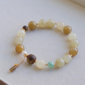 May include: A beaded bracelet featuring round polished stones in shades of cream, honey yellow, and light green, interspersed with small gold-toned spacers. A dark brown wooden bead, a white pearl, and a small gold chain dangle with a rectangular charm.
