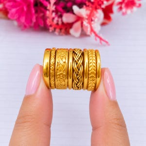Può includere: Un set di sei anelli color oro con varie texture e motivi, tra cui una fascia liscia, un design geometrico a rombi, un motivo floreale, uno stile a treccia, una fascia semplice e un motivo a foglie disposti in un'unica fila.