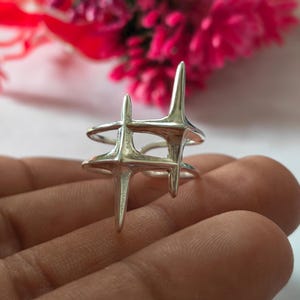 May include: A polished silver ring featuring two interconnected four-pointed star shapes. The design consists of two horizontal bands that meet at the star points, creating an open, geometric structure with a smooth, reflective metallic finish.
