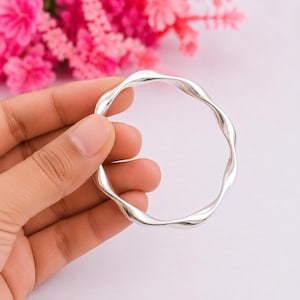 May include: A polished silver-tone bangle bracelet featuring a continuous, undulating wavy design. The circular band has a smooth, reflective metallic surface that catches the light, creating subtle highlights along its curved, rhythmic form.