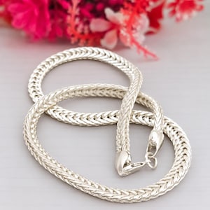 Op de afbeelding: Een zilverkleurige ketting met een dicht, gevlochten tarwepatroon. De ketting is afgewerkt met een gepolijste metallic glans en is voorzien van een veilige karabijnsluiting aan het uiteinde, wat zorgt voor een doorlopend en uniform ontwerp.