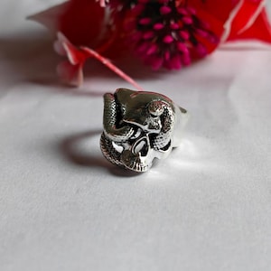 May include: A silver-coloured metal ring featuring a detailed skull design with a textured snake coiled around the cranium. The ring has a polished finish with darkened recessed areas to highlight the intricate features of the skull and the snake scales.