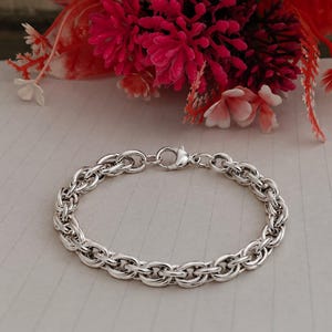 Può includere: Un bracciale a catena color argento caratterizzato da una serie di anelli metallici circolari intrecciati. Il bracciale è fissato con un piccolo meccanismo di chiusura a moschettone lucido, creando un design circolare continuo.