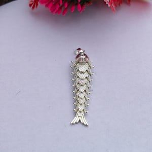 Pode incluir: Um pingente de peixe em tom prateado com um corpo segmentado e articulado, apresentando um padrão de escamas texturizado. O pingente possui uma pequena argola no topo para fixação e uma barbatana caudal detalhada na parte inferior do objeto.