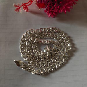 Peut inclure: Une chaîne de cou de type gourmette argentée composée de maillons plats et ovales entrelacés avec une finition polie et réfléchissante. La chaîne est enroulée en cercle et comporte un fermoir mousqueton sécurisé à l'une de ses extrémités.