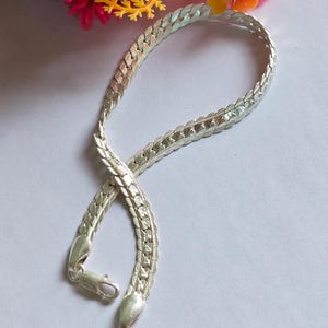 Könnte beinhalten: Ein silberfarbenes Kettenarmband mit einem flachen, geflochtenen Fischgrätenmuster und einer polierten Oberfläche. Das Armband verfügt über einen sicheren Karabinerverschluss an einem Ende und zeigt eine reflektierende metallische Textur.