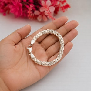 Può includere: Un bracciale color argento caratterizzato da un complesso motivo a maglia bizantina intrecciata. Il bracciale è composto da numerosi piccoli anelli metallici circolari intrecciati ed è fissato con una chiusura a moschettone lucida.