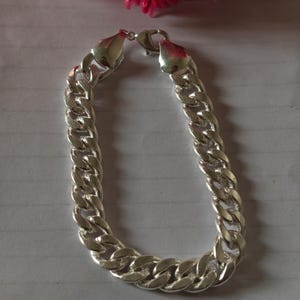 Könnte beinhalten: Ein silberfarbenes Panzerkettenarmband mit dicken, ineinandergreifenden ovalen Gliedern und einer polierten, reflektierenden Oberfläche. Das Armband wird durch einen Karabinerverschluss an der Oberseite geschlossen, der die Enden verbindet.