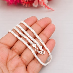 Könnte beinhalten: Eine silberfarbene Schlangenkette mit einer glatten, flexiblen und eng gewebten metallischen Textur. Die Kette verfügt über einen polierten Karabinerverschluss an den Enden, der dazu dient, die Kette zu einer durchgehenden Schlaufe zu schließen.