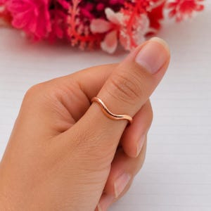 May include: A rose gold ring with a smooth, polished finish and a single gentle wave-like curve along the band. The metal band has a rounded profile and a consistent thickness, encircling the thumb with a simple and clean design aesthetic.