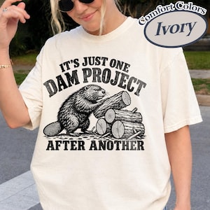 May include: An ivory short-sleeved t-shirt featuring a black graphic of a beaver stacking logs. The text reads IT'S JUST ONE DAM PROJECT AFTER ANOTHER. The shirt has a crew neckline and a relaxed fit, with a Comfort Colors Ivory label visible.