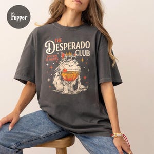 Könnte beinhalten: Ein anthrazitfarbenes T-Shirt mit einer Grafik einer Katze, die eine Sonnenbrille und eine Krone trägt und hinter einem Cocktailglas mit einer Kirsche sitzt. Der Text lautet THE DESPERADO CLUB und SO FUN IT HURTS, umgeben von Sternen.
