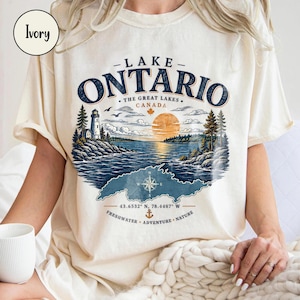 Könnte beinhalten: Ein elfenbeinfarbenes T-Shirt mit einer Grafik des Lake Ontario, die einen Leuchtturm, Kiefern, einen Sonnenuntergang und eine Windrose zeigt. Der Text lautet Lake Ontario, The Great Lakes, Canada, 43.6532 N, 78.4487 W, Freshwater, Adventure, Nature.