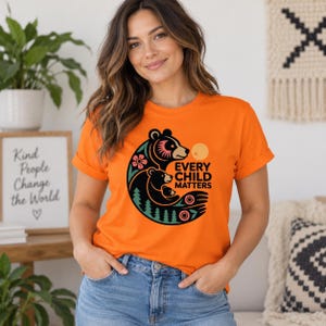 May include: An orange short-sleeve t-shirt featuring a graphic of three black bears in a circular arrangement with green pine trees, pink flowers, and a yellow sun. The text "EVERY CHILD MATTERS" is printed in black next to the bear graphic.