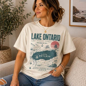 Könnte beinhalten: Ein cremefarbenes Kurzarm-T-Shirt mit einem grafischen Aufdruck einer Karte des Ontariosees. Das Design enthält den Text Lake Ontario, The Great Lakes, Freshwater Sea sowie verschiedene nautische Illustrationen.