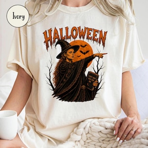 Könnte beinhalten: Ein elfenbeinfarbenes T-Shirt mit einer Grafik einer Hexe in schwarzem Umhang und spitzem Hut, die auf einen orangefarbenen Mond mit Fledermäusen zeigt. Der Text "HALLOWEEN" steht in Orange darüber, und die Hexe hält ein Buch "I DON'T HAVE PEN FOR ME".