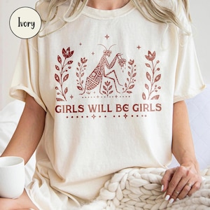 May include: An ivory short-sleeve t-shirt featuring a dark red graphic print of a praying mantis surrounded by floral branches and stars. The text GIRLS WILL BE GIRLS is printed in a bold, serif font below the central insect illustration.
