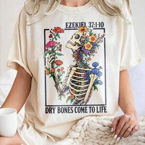 Peut inclure: Un t-shirt couleur crème avec une impression graphique représentant un squelette entouré de fleurs colorées. Le texte EZEKIEL 37:1-10 figure en haut et le texte DRY BONES COME TO LIFE est imprimé en bas, le tout dans un cadre noir.