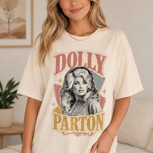 May include: An off-white short-sleeve t-shirt featuring a graphic print of Dolly Parton. The design includes the text DOLLY PARTON in bold lettering, surrounded by a distressed, multi-coloured background with star accents and a vintage aesthetic.