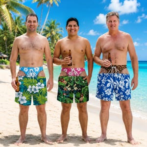 May include: Three pairs of swim trunks featuring tropical floral patterns in green, black, and blue. Each pair has a white drawstring waist and a high-cut leg design. The trunks are displayed on three individuals standing on a sandy beach.