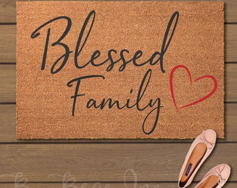 May include: A rectangular coir doormat with the words "Blessed Family" in black script and a red heart. Two pairs of pink ballet flats with bows are next to the doormat. The background is a wooden surface.