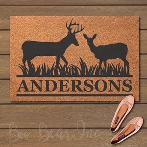 May include: A rectangular doormat with a deer silhouette design. The mat features a buck and a doe, with the family name "Andersons" in a bold font. The doormat is made of a natural fiber material and is placed on a wooden surface.
