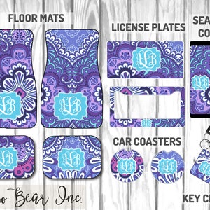 May include: A collection of car accessories with a purple, blue, and white paisley design. Items include floor mats, license plates, seat belt covers, car coasters, and a keychain. Each item features a light blue monogram. The text "Boo Bear Inc." is visible.