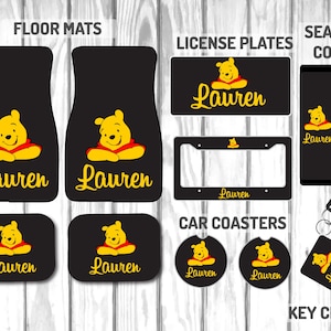 May include: A range of car accessories with a yellow cartoon bear in a red shirt and the name "Lauren" in gold script. The items include floor mats, license plates, seat belt covers, car coasters, and a keychain, all on a black background.