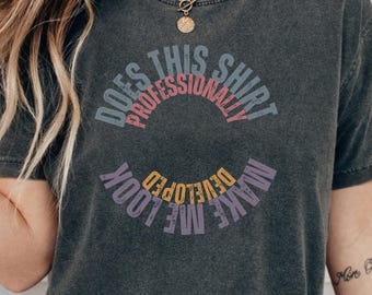 May include: A charcoal gray short-sleeve t-shirt featuring circular text that reads Does This Shirt Make Me Look Professionally Developed in muted blue, pink, purple, and yellow lettering across the center of the chest area.