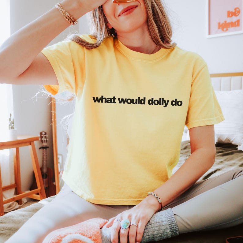 May include: A bright yellow short-sleeved t-shirt featuring the black text "what would dolly do" printed across the chest. The garment has a crew neckline and a relaxed fit, constructed from a solid-colored fabric with a smooth texture.