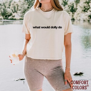 May include: A cream-colored short-sleeve t-shirt featuring the black text what would dolly do printed across the chest. The shirt has a crew neckline and a relaxed fit, paired with ribbed mauve-colored shorts that reach mid-thigh.