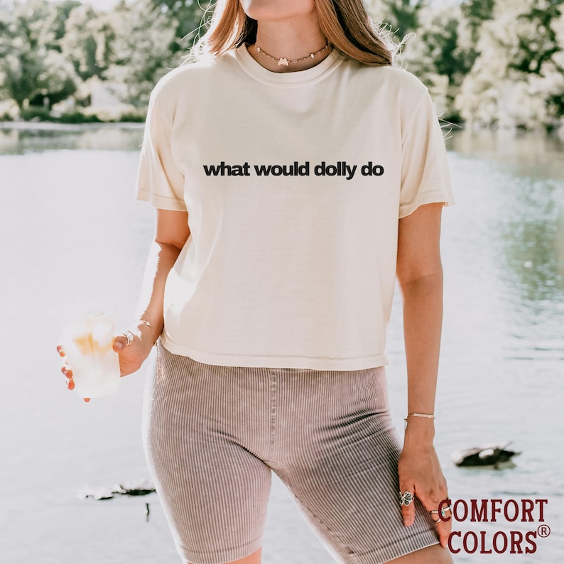May include: A cream-colored short-sleeve t-shirt featuring the black text what would dolly do printed across the chest. The shirt has a crew neckline and a relaxed fit, paired with ribbed mauve-colored shorts that reach mid-thigh.
