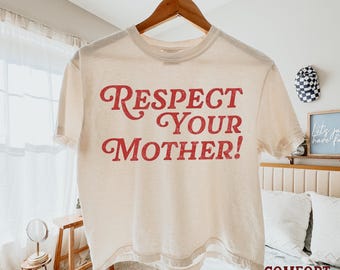 May include: A cream-colored short-sleeve t-shirt featuring the red text "RESPECT YOUR MOTHER!" printed in a bold, distressed font across the chest. The garment is displayed on a wooden hanger against a bright, neutral-toned indoor setting.