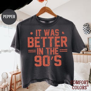 May include: A charcoal gray short-sleeve t-shirt hanging on a wooden hanger. The shirt features the orange distressed text "IT WAS BETTER IN THE 90'S" printed on the front. The fabric has a washed, vintage appearance with a crew neckline.