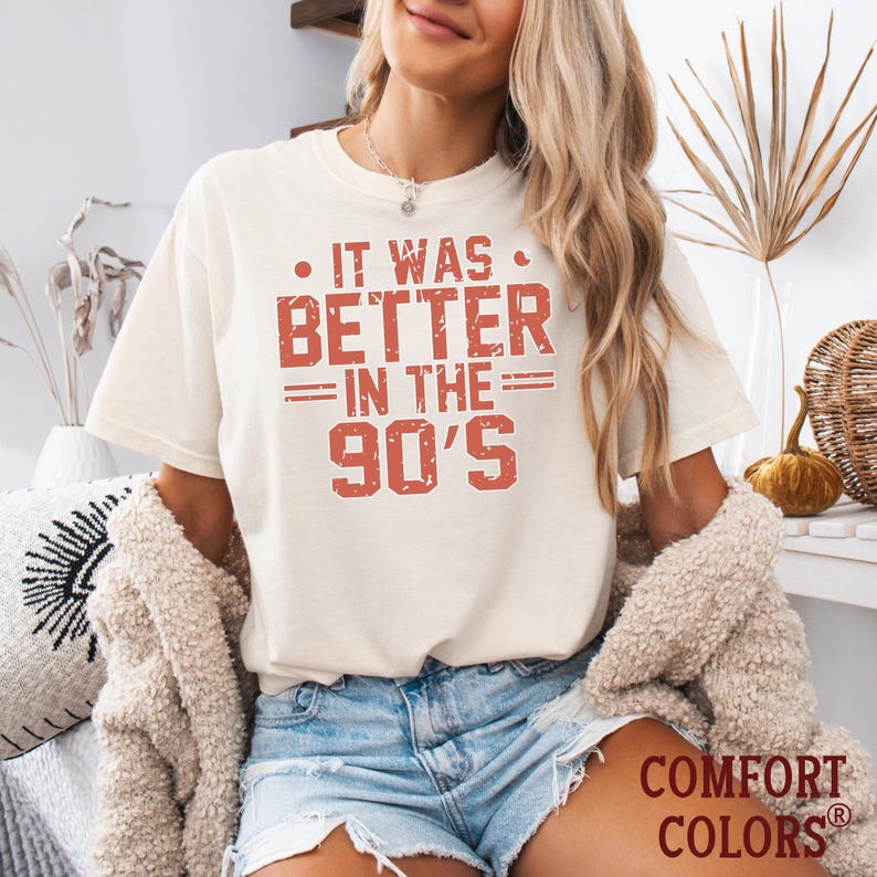 May include: A cream-colored short-sleeve t-shirt featuring the distressed red text IT WAS BETTER IN THE 90'S printed on the front. The shirt has a crew neckline and a relaxed fit, with the Comfort Colors brand logo visible in the bottom corner.