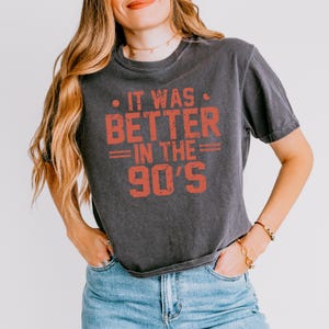May include: A charcoal grey short-sleeve t-shirt featuring the distressed orange text "IT WAS BETTER IN THE 90'S" printed on the front. The shirt has a crew neckline and a relaxed fit, paired with blue denim jeans.