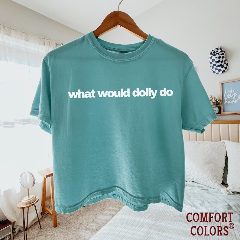 May include: A teal short-sleeve crew neck t-shirt featuring the white text "what would dolly do" printed across the chest. The garment is displayed on a wooden hanger and has a solid color finish with a standard fit and hemmed sleeves.