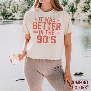 May include: A cream-colored short-sleeve t-shirt featuring the distressed red text IT WAS BETTER IN THE 90'S printed on the front. The shirt has a crew neckline and a relaxed fit, paired with ribbed, fitted shorts in a muted mauve-brown color.