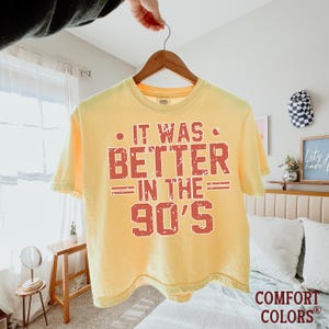 May include: A pale yellow short-sleeve t-shirt with a distressed red graphic print on the front that reads IT WAS BETTER IN THE 90'S. The shirt features a crew neckline and is displayed on a wooden hanger against a bright indoor setting.
