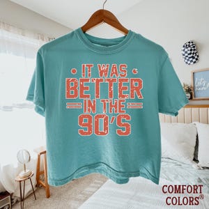 May include: A teal short-sleeved t-shirt with a crew neckline. The front features distressed, block-style red text that reads IT WAS BETTER IN THE 90'S, with horizontal lines flanking the word IN. The shirt is displayed on a wooden hanger.