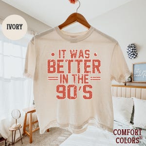 May include: An ivory short-sleeve t-shirt featuring the red distressed text IT WAS BETTER IN THE 90'S centered on the chest. The shirt is displayed on a wooden hanger against a light-colored background. The fabric has a soft, lived-in appearance.