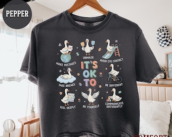 May include: A charcoal gray t-shirt featuring a central graphic with the text "IT'S OK TO" surrounded by illustrations of white geese. The geese are depicted in various activities with labels including "MAKE MISTAKES," "UNMASK," "AVOID EYE CONTACT," "TAKE BREAKS," "BE DIFFERENT," "FEEL DEEPLY," "BE YOURSELF," and "COMMUNICATE DIFFERENTLY."