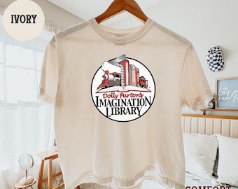 May include: An ivory t-shirt featuring a circular graphic of a red train engine emerging from a stack of books. The text reads Dolly Parton's IMAGINATION LIBRARY. The shirt is displayed on a wooden hanger against a light-colored bedroom background.