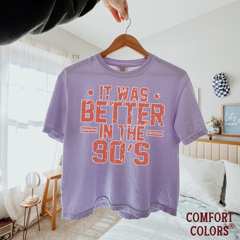 May include: A light purple short-sleeve t-shirt featuring the text IT WAS BETTER IN THE 90'S in a distressed orange font. The shirt has a crew neckline and is displayed on a wooden hanger against a bright, indoor bedroom setting.