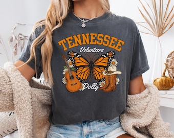 May include: A charcoal gray short-sleeve t-shirt featuring a graphic print with the text "TENNESSEE Volunteers Dolly". The design includes a large orange and black monarch butterfly, an acoustic guitar, cowboy boots, and white daisies.