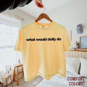 May include: A pale yellow short-sleeve t-shirt featuring the black text "what would dolly do" printed across the chest. The garment has a crew neckline and is displayed on a wooden hanger, showcasing a simple design with a solid color base.