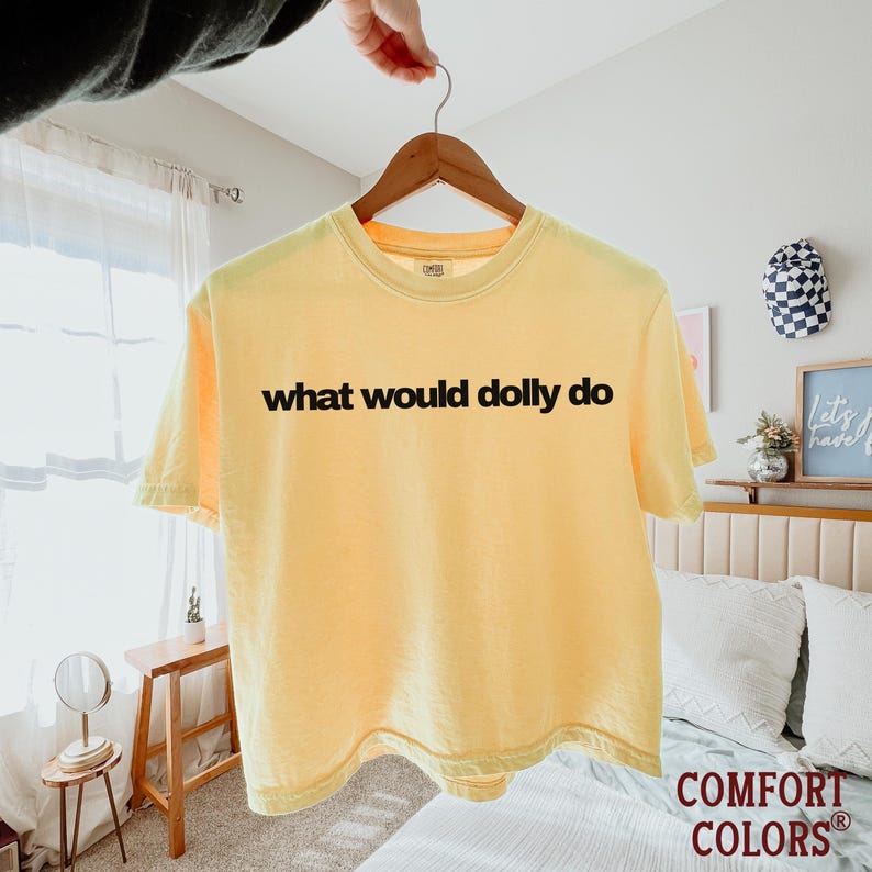 May include: A pale yellow short-sleeve t-shirt featuring the black text "what would dolly do" printed across the chest. The garment has a crew neckline and is displayed on a wooden hanger, showcasing a simple design with a solid color base.