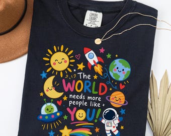May include: A black t-shirt featuring a colorful space-themed graphic with a sun, moon, rocket, planet, and astronaut. The text reads, The WORLD needs more people like YOU! The design includes stars, hearts, and a UFO, all in vibrant colors.